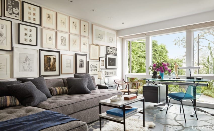Modern living room with gray sofa, art-covered walls, and large window offering garden view. Desk and chair near window.