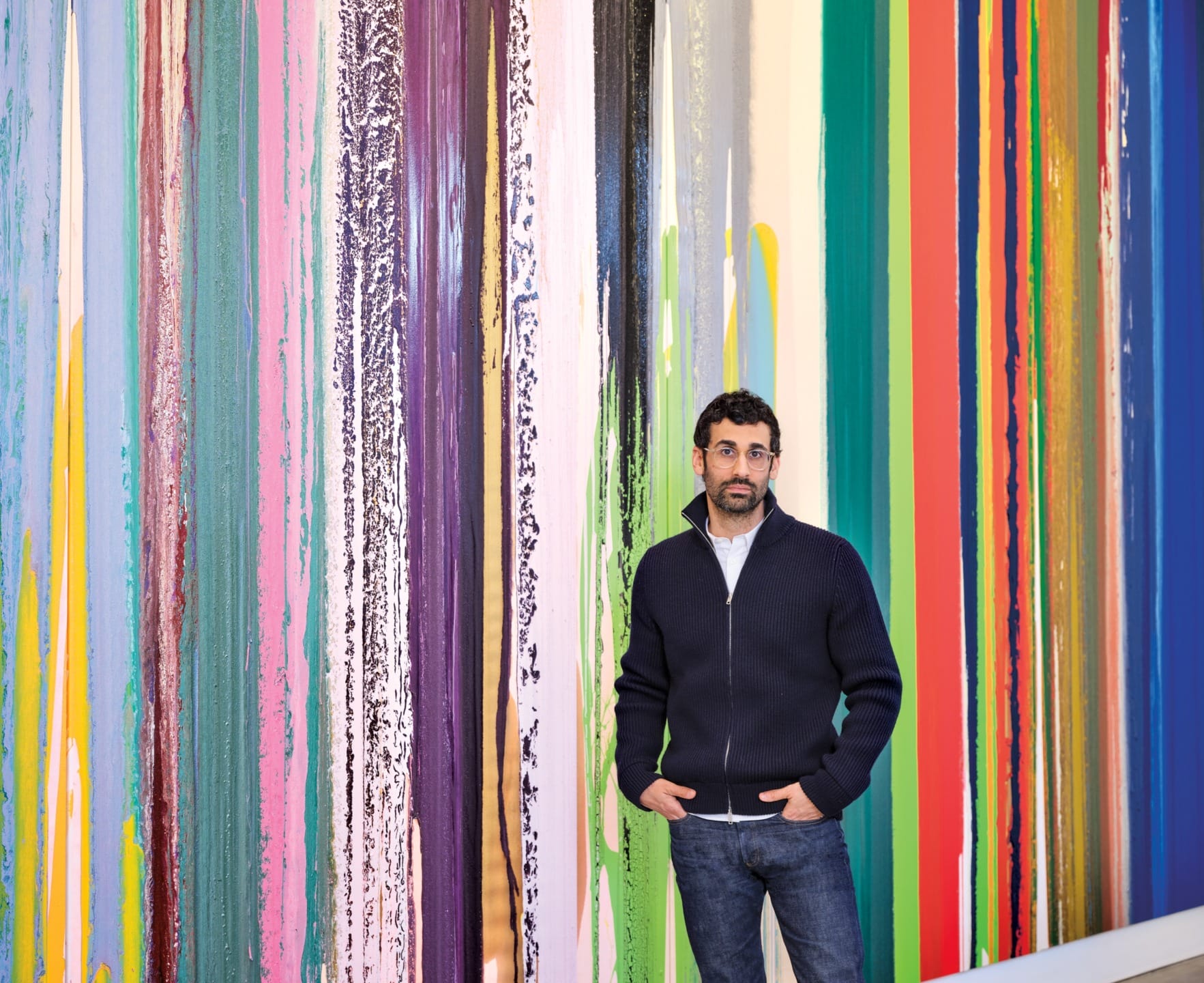 Man standing in front of a colorful wall with vertical paint drips, wearing a dark sweater and jeans.