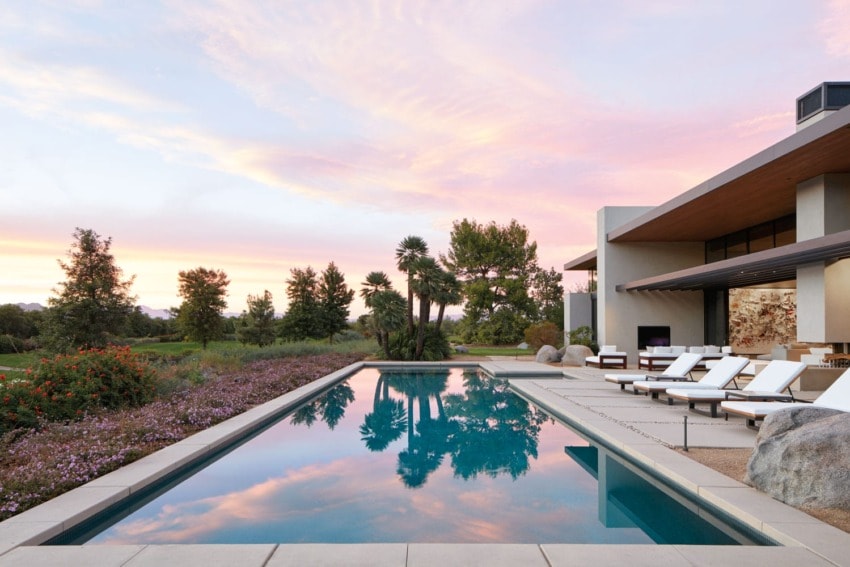 Modern house with pool at sunset, lounge chairs aligned on patio, trees in the background, sky with pink and orange clouds