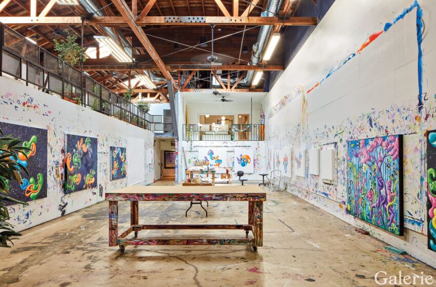 Colorful art studio with high ceilings, wooden beams, large canvas paintings on walls, and a central workspace table.
