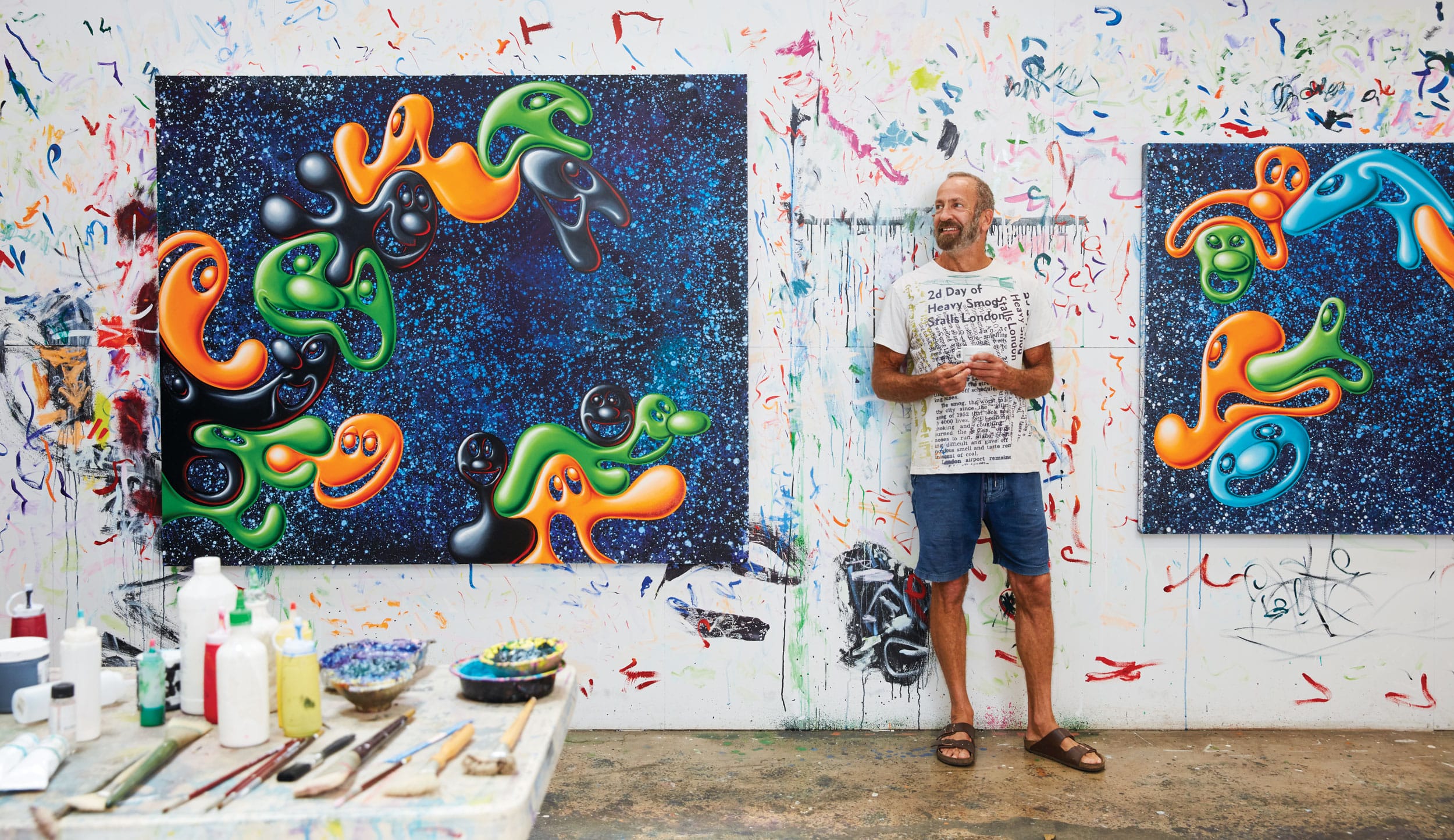 Artist stands beside vibrant abstract paintings in a colorful studio, surrounded by scattered art supplies and a paint-splattered wall.