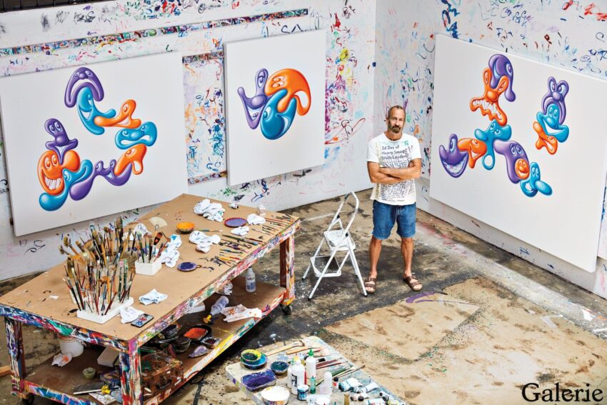 Artist standing in a colorful studio with abstract paintings, paint splatters on walls, and art supplies on a table.