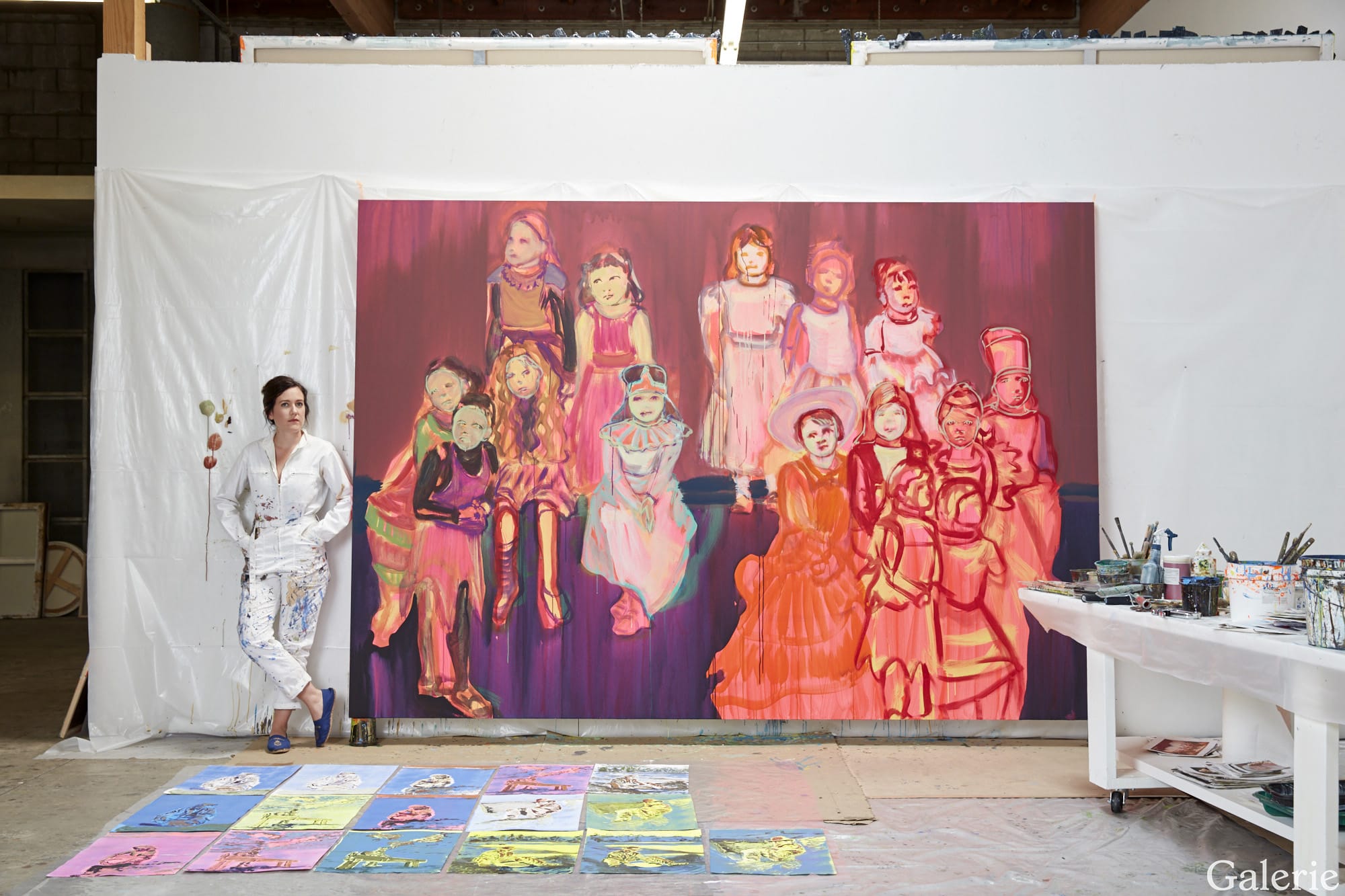 Artist in studio with a large colorful painting of figures; painting supplies and canvas visible.