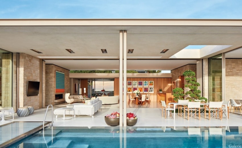 Modern open-air living space with a pool, contemporary furniture, artwork, and potted plants under a clear blue sky.