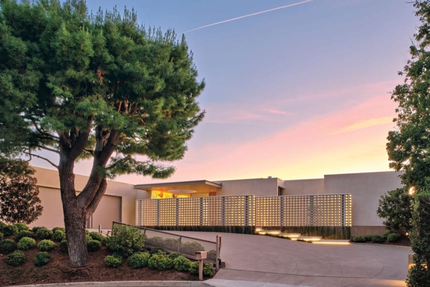 Modern house exterior at sunset with a large tree, unique architectural design, and a clear sky in the background.