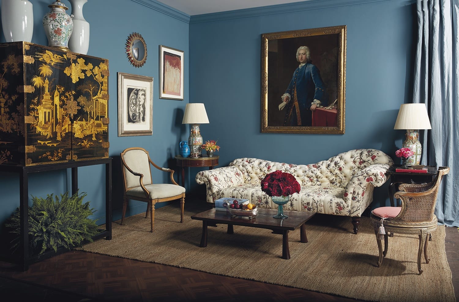 Eclectic living room with blue walls, floral sofa, various chairs, antique cabinet, and framed artwork.