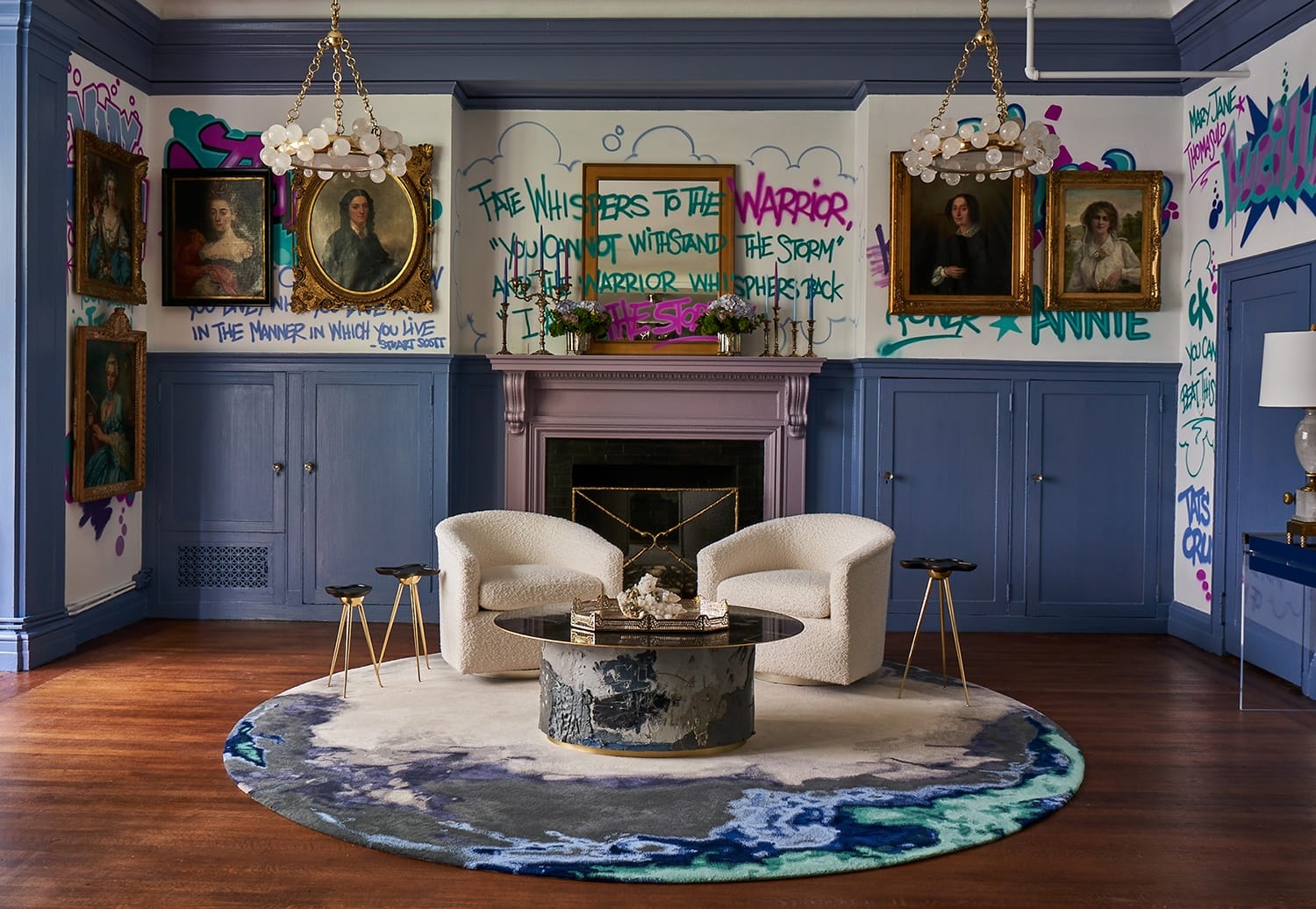 Eclectic living room with graffiti walls, elegant chandeliers, portraits, white chairs, and a round table on a patterned rug.