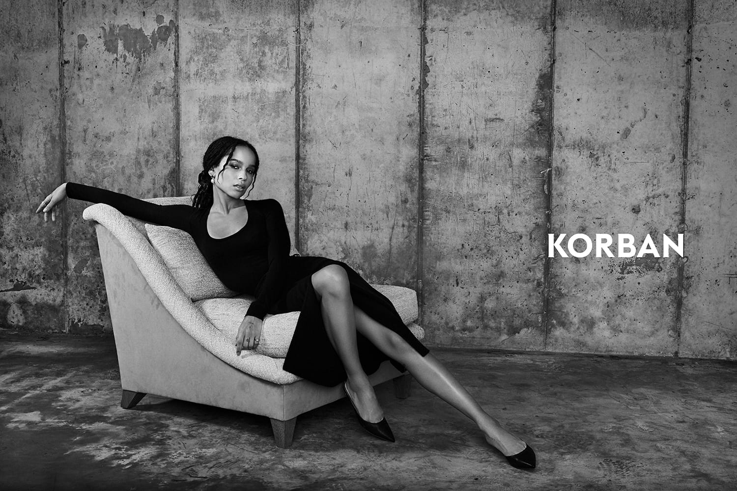 Person in a black dress lounging on a chair against a concrete wall background with the text "KORBAN" beside them.