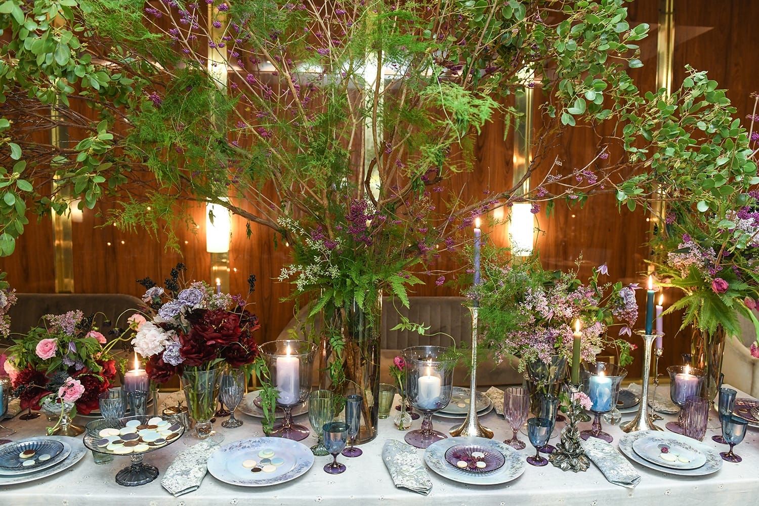 Elegant dining table setting with lush greenery, flowers, candles, and decorative plates in a vibrant, opulent atmosphere.