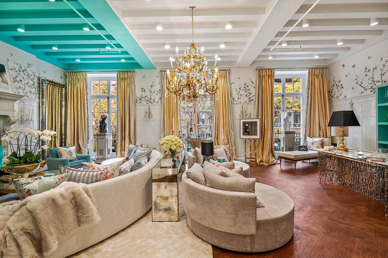Luxurious living room with elegant chandelier, curved sofas, decorative wallpaper, and large windows with gold curtains.