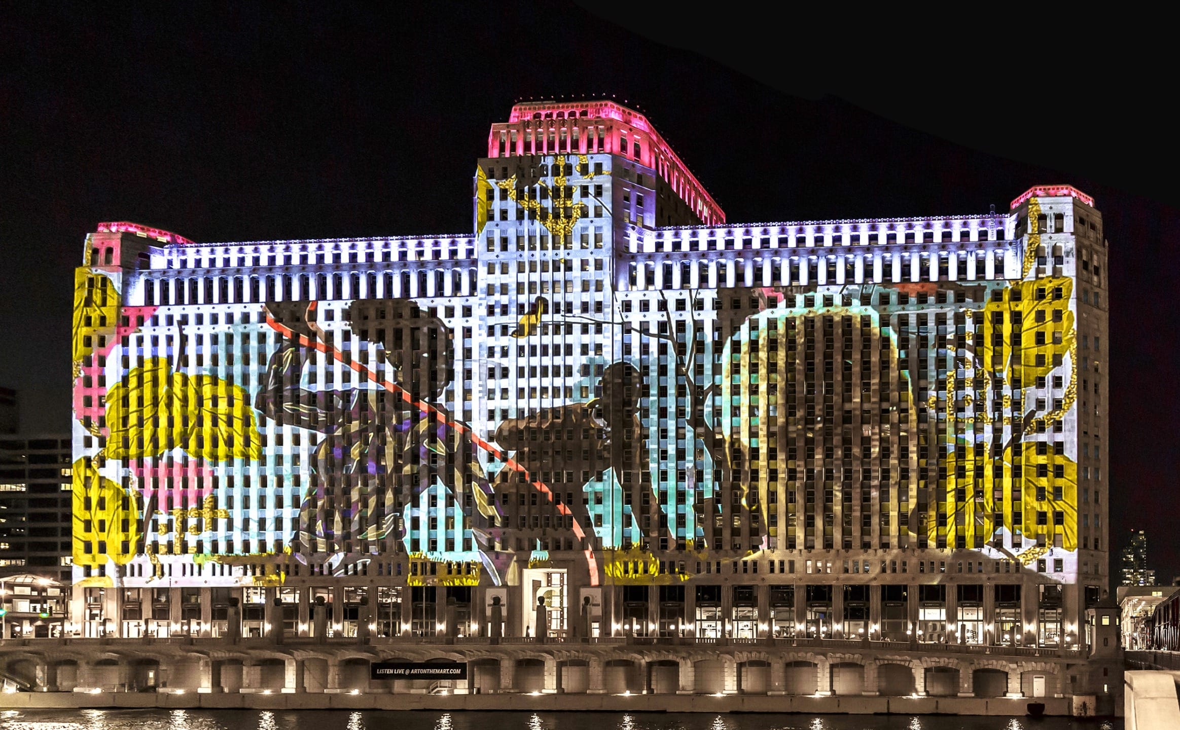 Colorful projection of artistic figures on an illuminated building at night along the river.