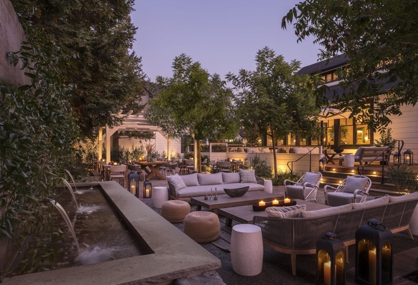 Cozy outdoor patio with sofas, coffee tables, soft lighting, lush trees, and a water feature at dusk.