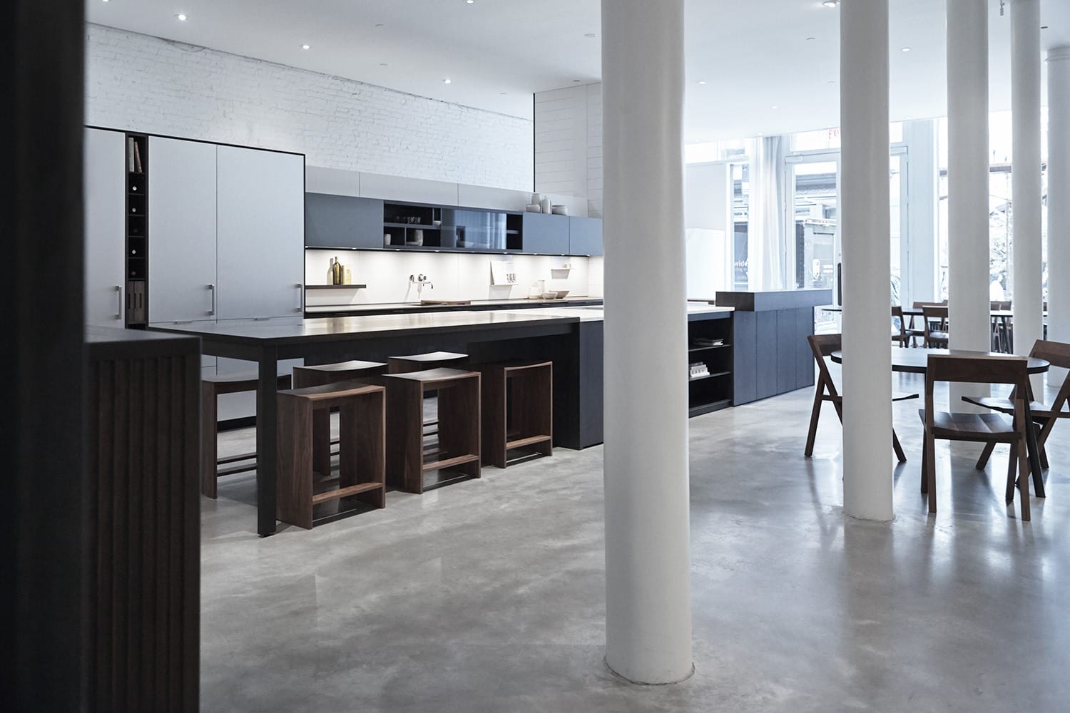 Modern kitchen with sleek countertops, wooden stools, and natural light streaming through large windows.