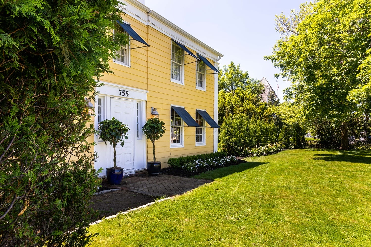 Yellow house with green shutters and large front lawn surrounded by trees and shrubs on a sunny day.