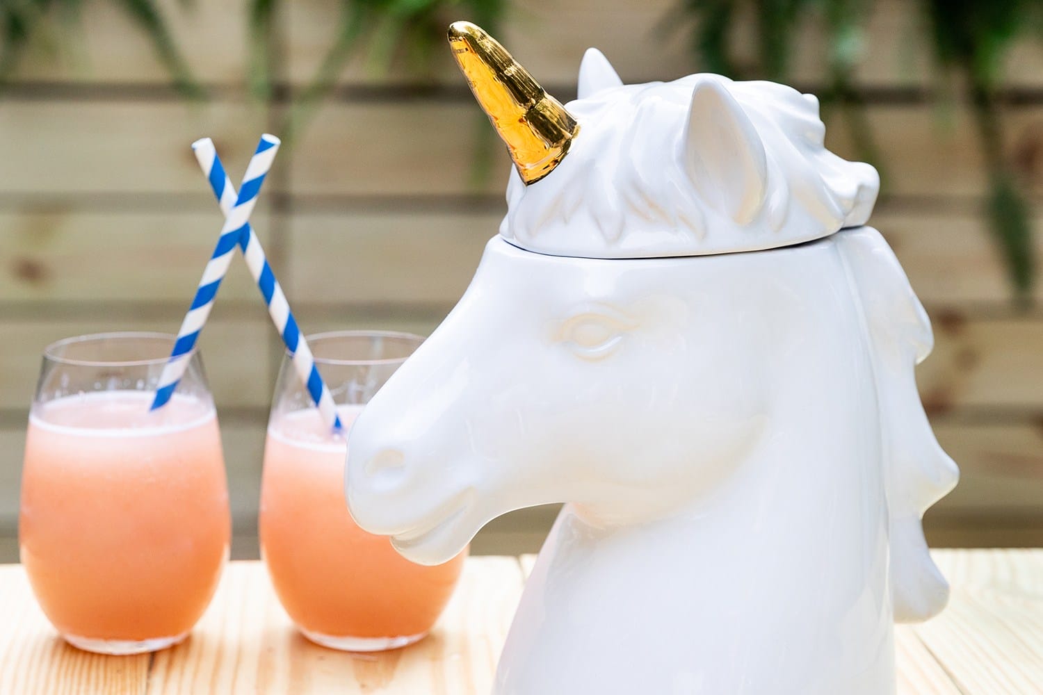 Ceramic unicorn head with golden horn beside two pink smoothies on a wooden table with blue and white straws.