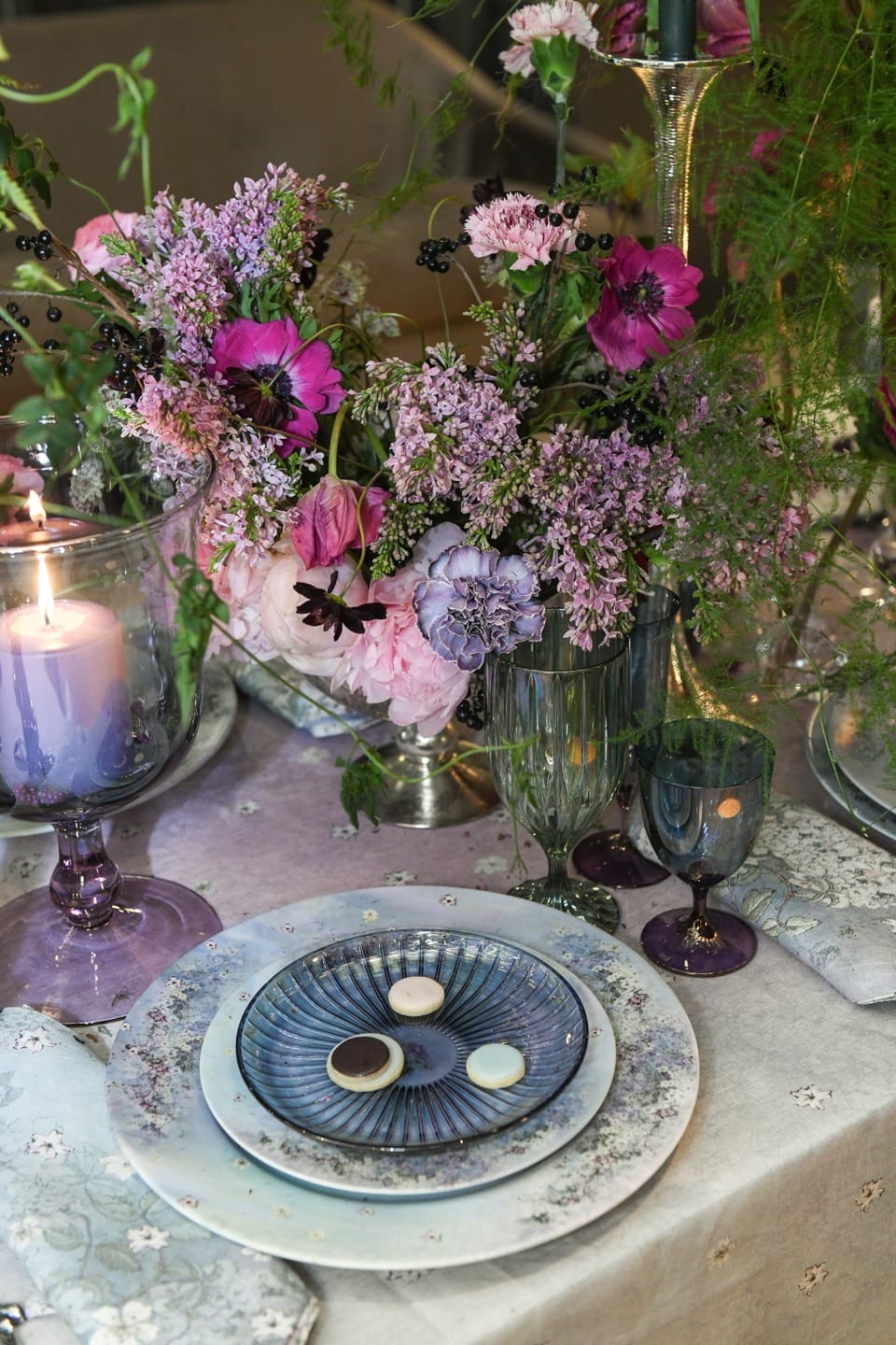 Elegant table setting with floral arrangement, purple candle, blue patterned plates, and glasses on a decorated table.