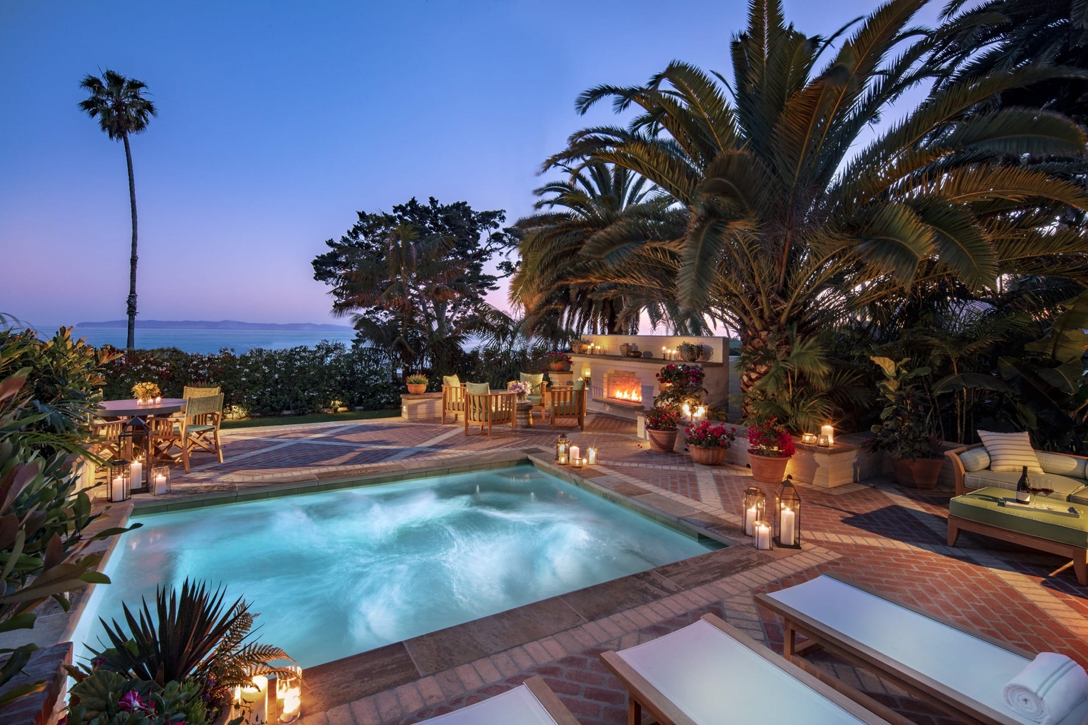Luxurious backyard with illuminated pool, cozy seating, palm trees, and a fireplace at dusk.