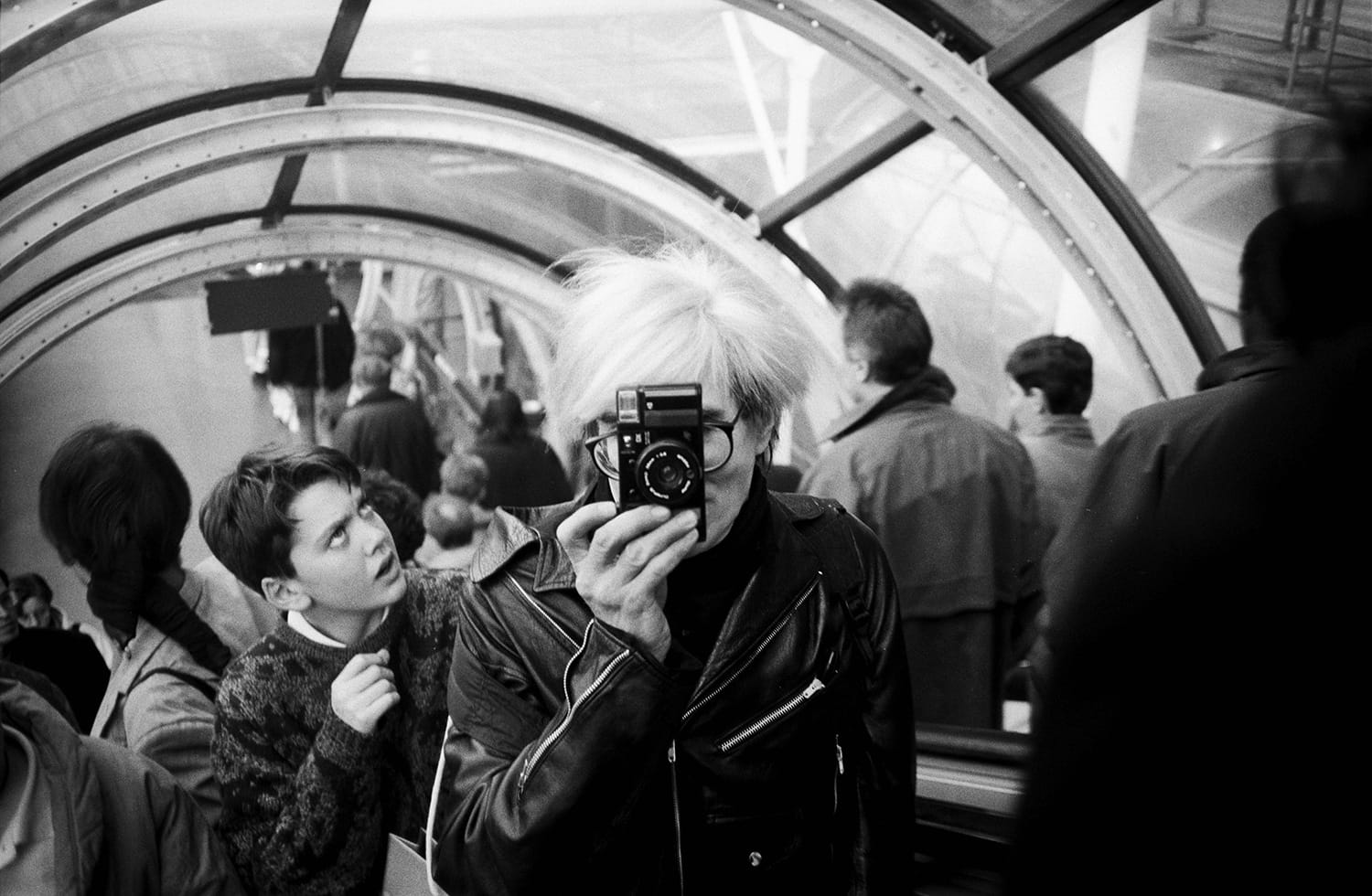 Person with white hair taking a photo in a crowded, glass-covered escalator.