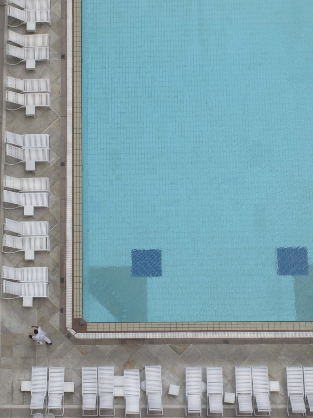 Aerial view of an empty swimming pool with white lounge chairs lined up on the deck and a person walking by.