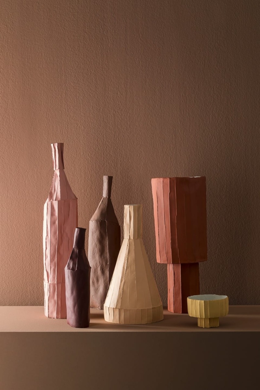 Assorted geometric ceramic vases and a bowl in earthy tones on a beige surface against a brown textured wall.