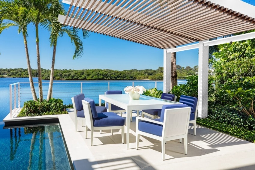 Outdoor patio with a white dining set and blue cushions by a lake under a pergola, surrounded by lush greenery and palm trees.