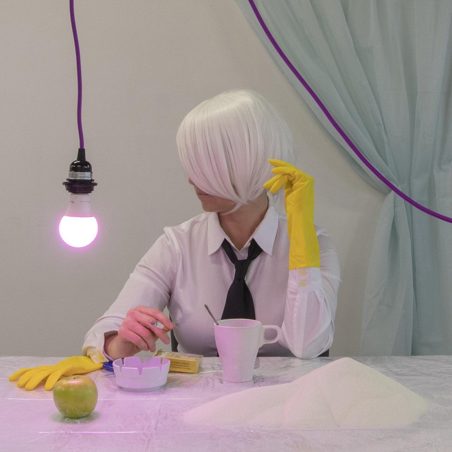 Person with short white hair and yellow gloves sitting at a table with a green apple, cup, cigar, and sugar pile.