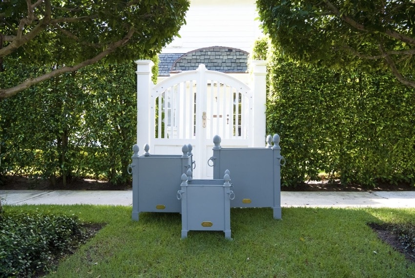White garden gate framed by green hedges with three gray planter boxes on a well-maintained lawn.