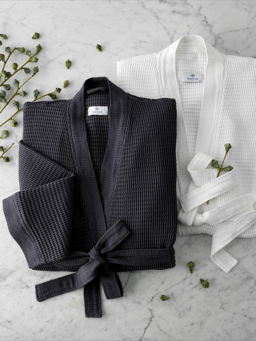Two folded waffle-knit bathrobes, one black and one white, on a marble surface with scattered green leaves.
