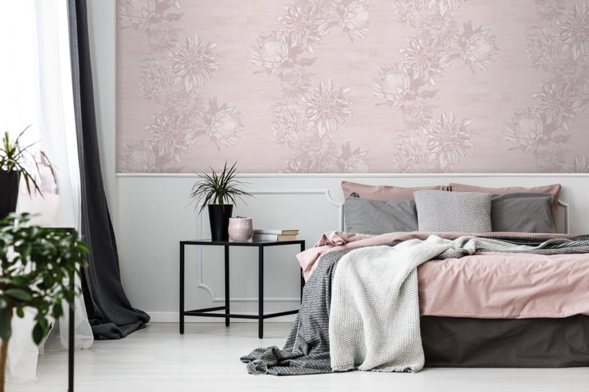 Cozy bedroom with floral patterned wall, pink and gray bedding, small black table with plants, and white draped curtains.