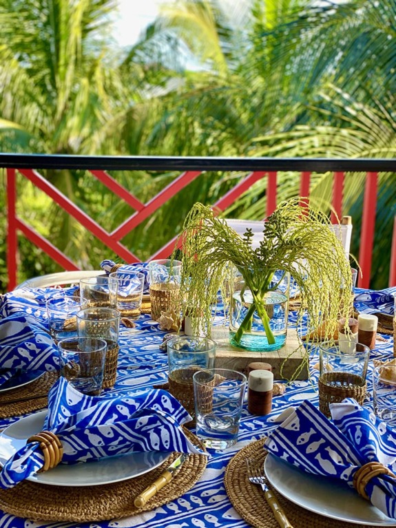 Outdoor dining table with blue patterned napkins, glassware, and greenery centerpiece on a balcony amidst palm trees.