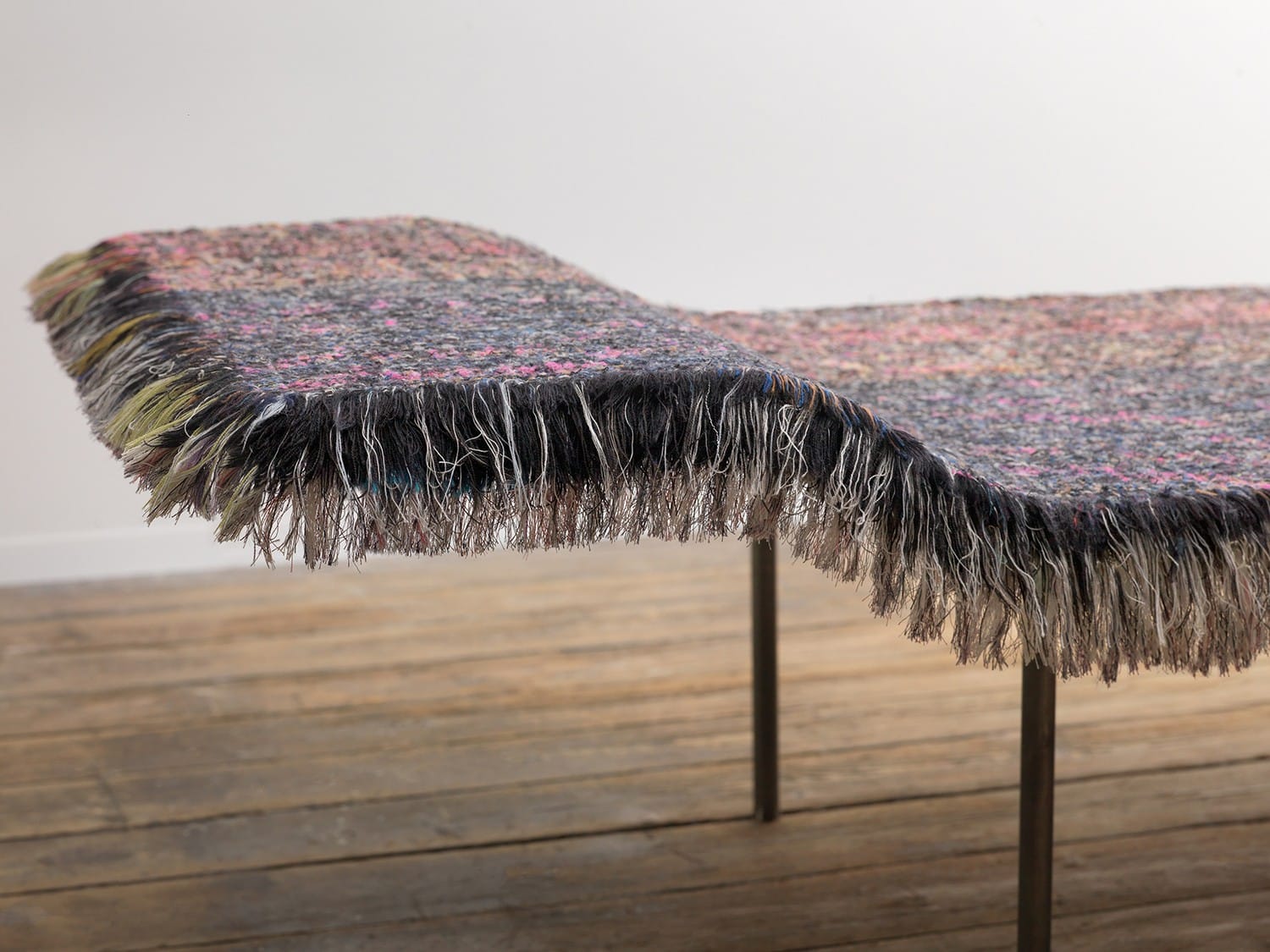 Fringed woven textile art hanging on metal stand on wooden floor in a minimalist room.