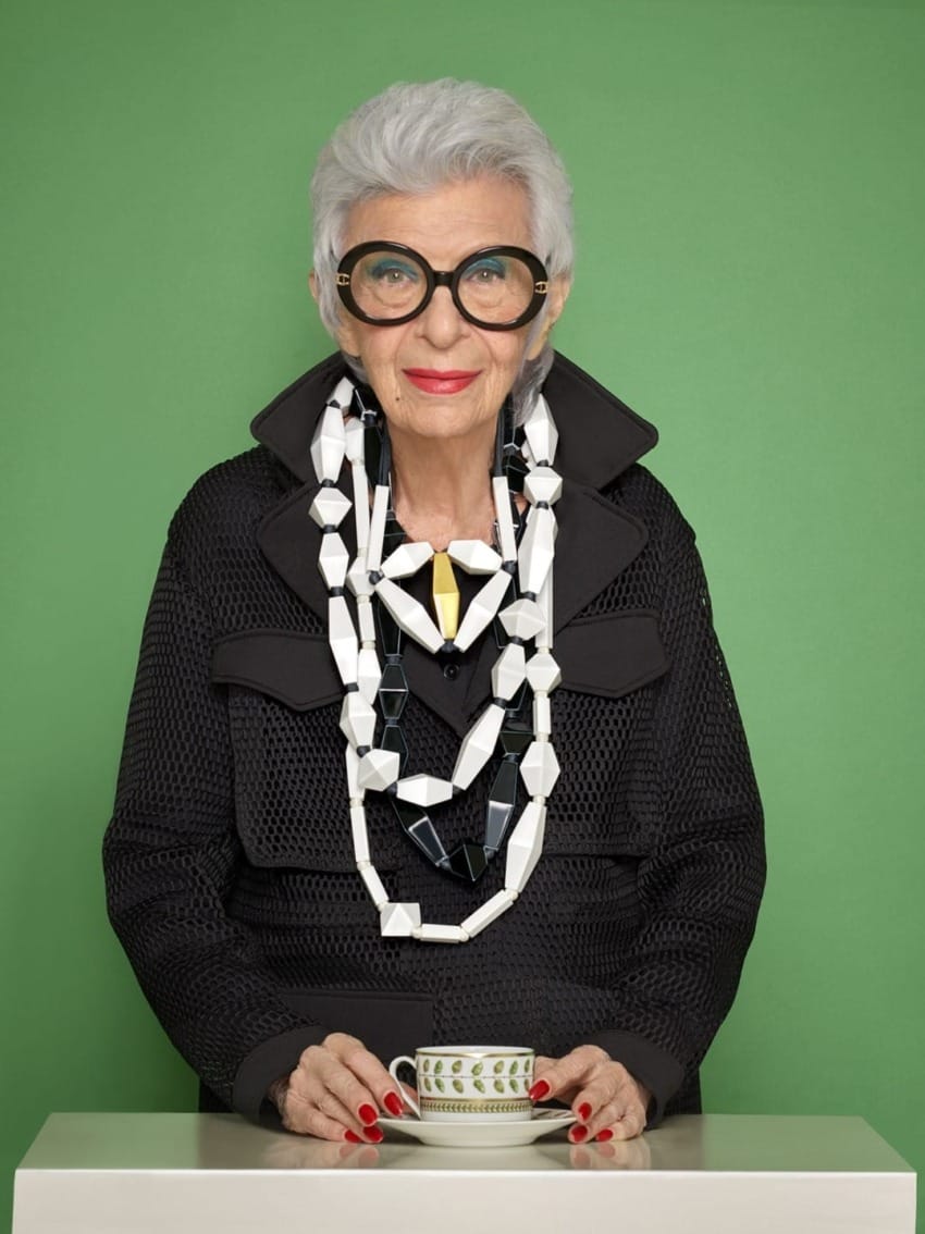 Elderly woman with short white hair, wearing large glasses and chunky jewelry, holds a patterned teacup against a green background.