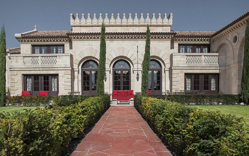 Elegant stone mansion with arched windows, manicured hedges, and a red pathway leading to the entrance.