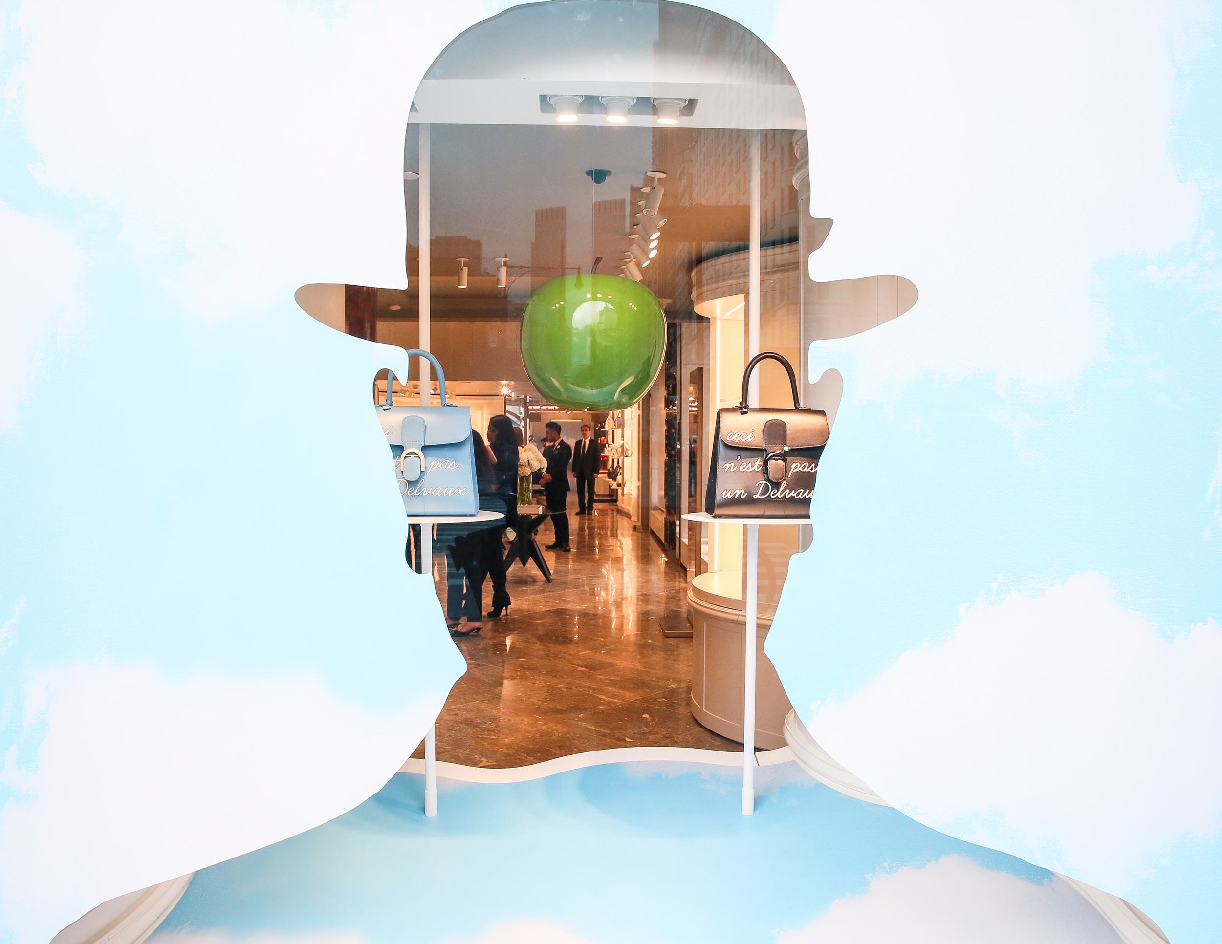 Storefront window display with a surreal silhouette, an apple, and bags against a blue sky background.