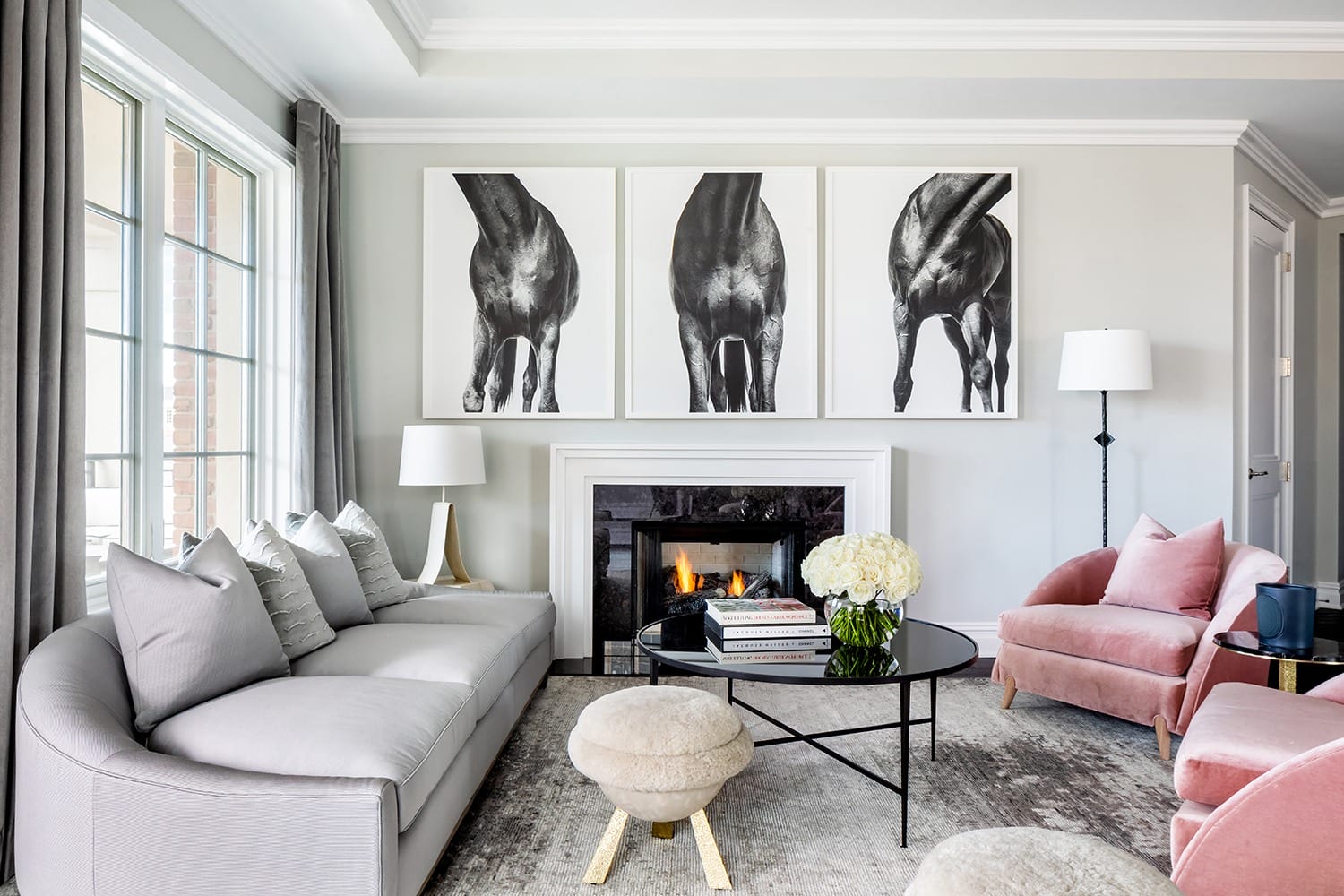 Contemporary living room with gray sofa, pink chairs, a round coffee table, and a fireplace under horse artwork.