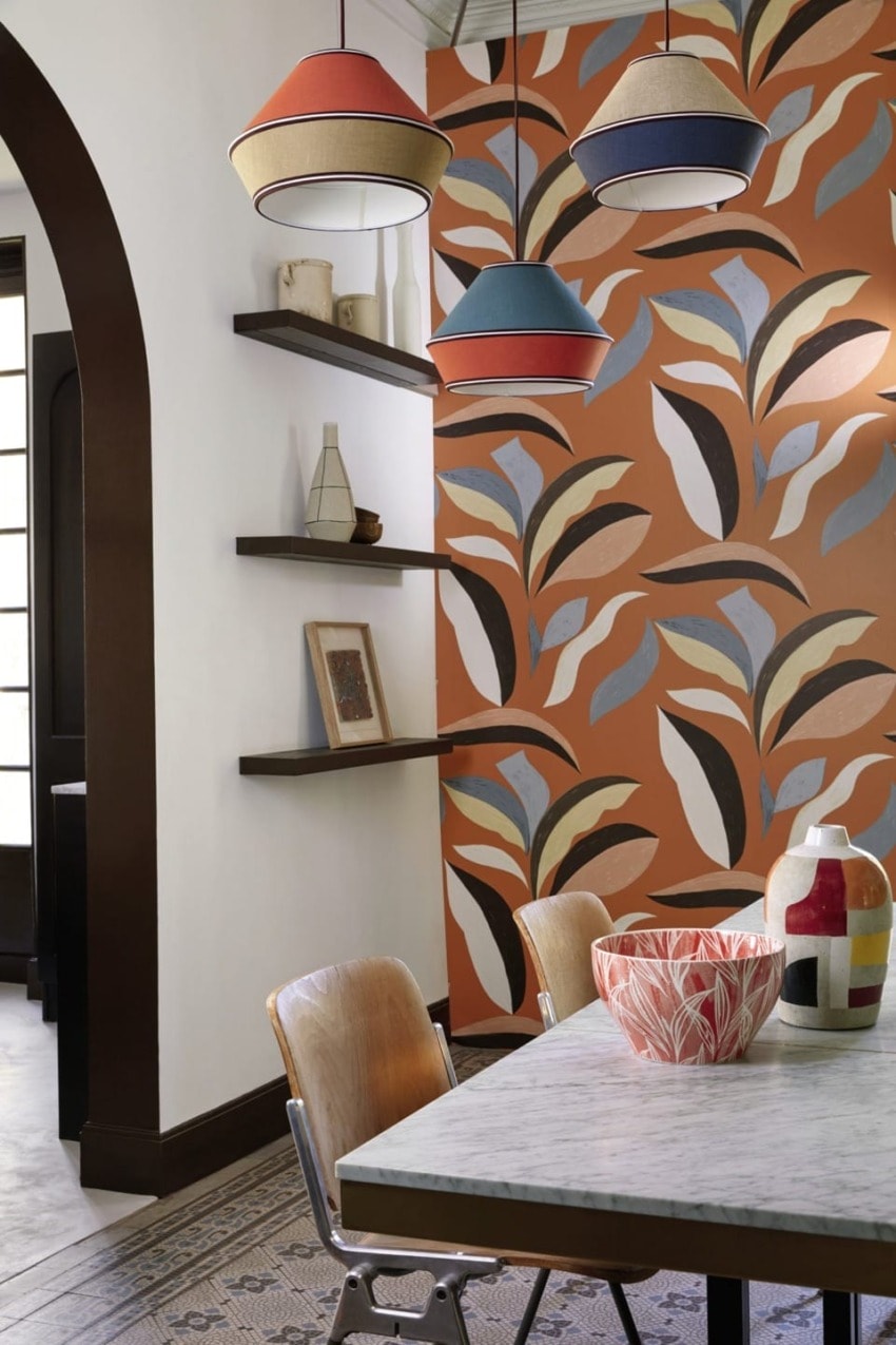 Dining area with marble table, wooden chairs, colorful pendant lights, decorative vases, and bold leaf-patterned wallpaper.