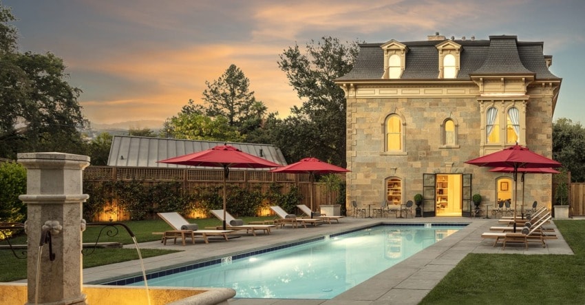 Luxurious stone villa with a pool, red umbrellas, sun loungers, and a landscaped garden at sunset.