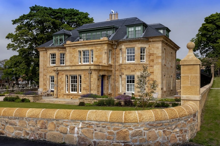 Large stone house with multiple windows, a slate roof, and a surrounding stone wall, set against a backdrop of trees and sky.