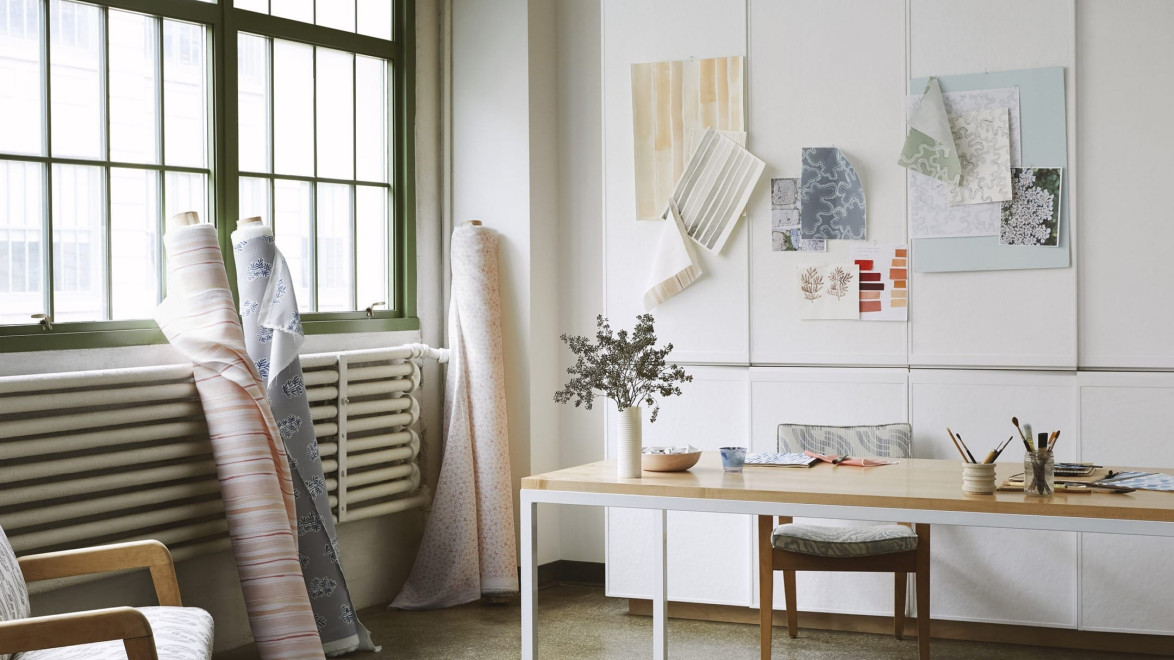 Interior design studio with fabric samples on walls, rolls of fabric by the window, and a wooden table with design materials.