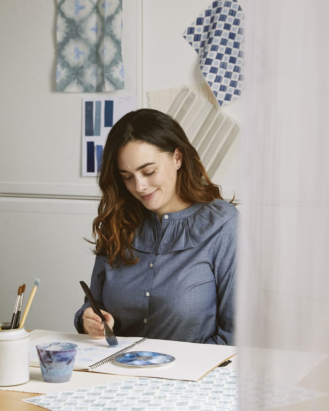Woman painting in a studio with patterned fabric swatches displayed on the wall in the background.