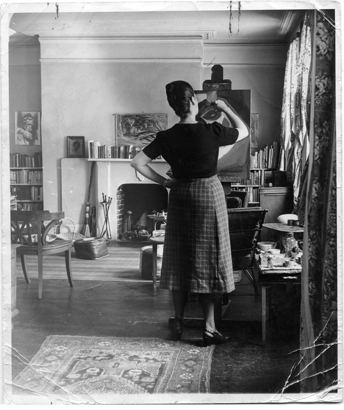 A woman stands in a vintage living room, painting at an easel with bookshelves and a fireplace in the background.