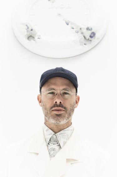 A person wearing a white coat, patterned scarf, blue cap, and glasses, standing in front of a white wall clock.