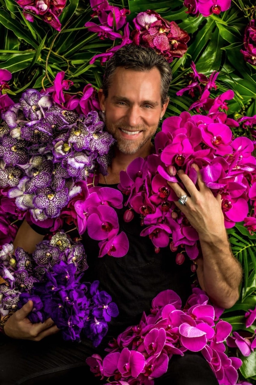 Man smiling while surrounded by vibrant pink and purple orchids, wearing a black shirt, holding flowers with green leaves in background.