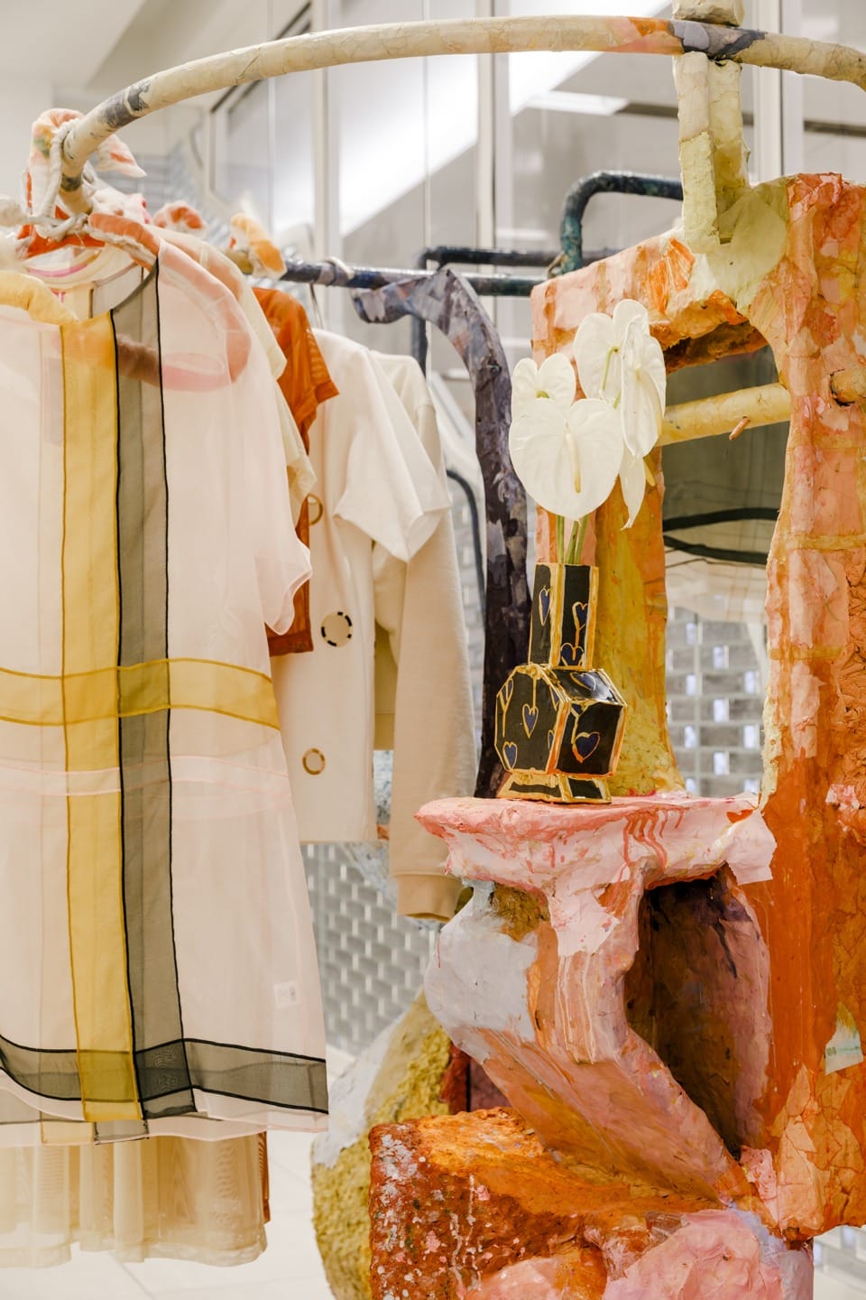 Art installation featuring colorful clothing and abstract sculptures, including a mirror and floral decor, in a lively display.