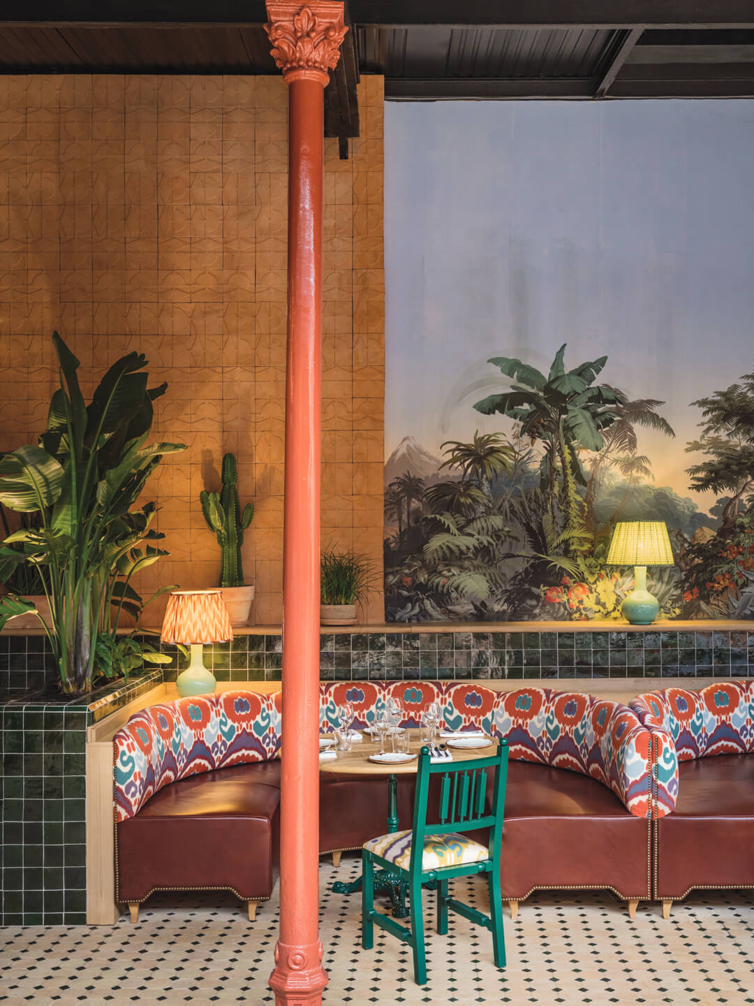 Colorful restaurant interior with patterned seating, tropical wallpaper, green chair, plants, red column, and dim lamps.