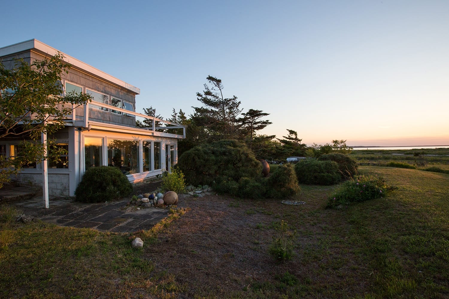 Modern house with large windows reflecting sunset, surrounded by lush greenery and overlooking a scenic coastal landscape.