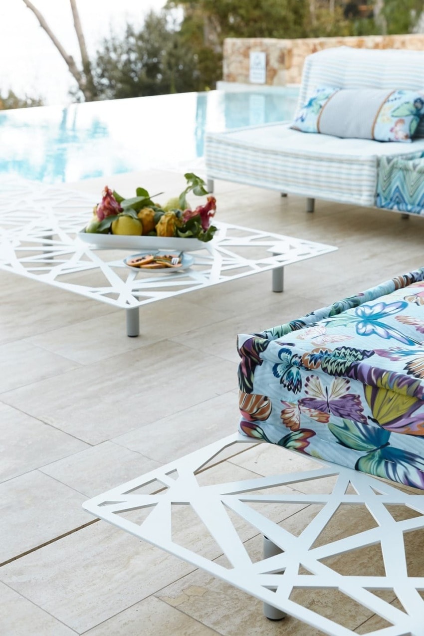 Poolside lounge area with modern white geometric tables, tropical floral cushions, and fruit platter.