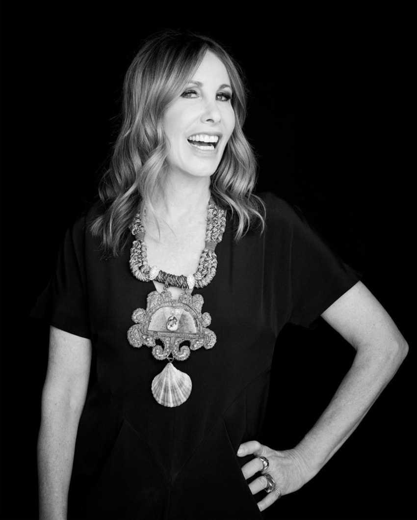 Smiling woman with wavy hair wearing an ornate necklace and black dress against a dark background.