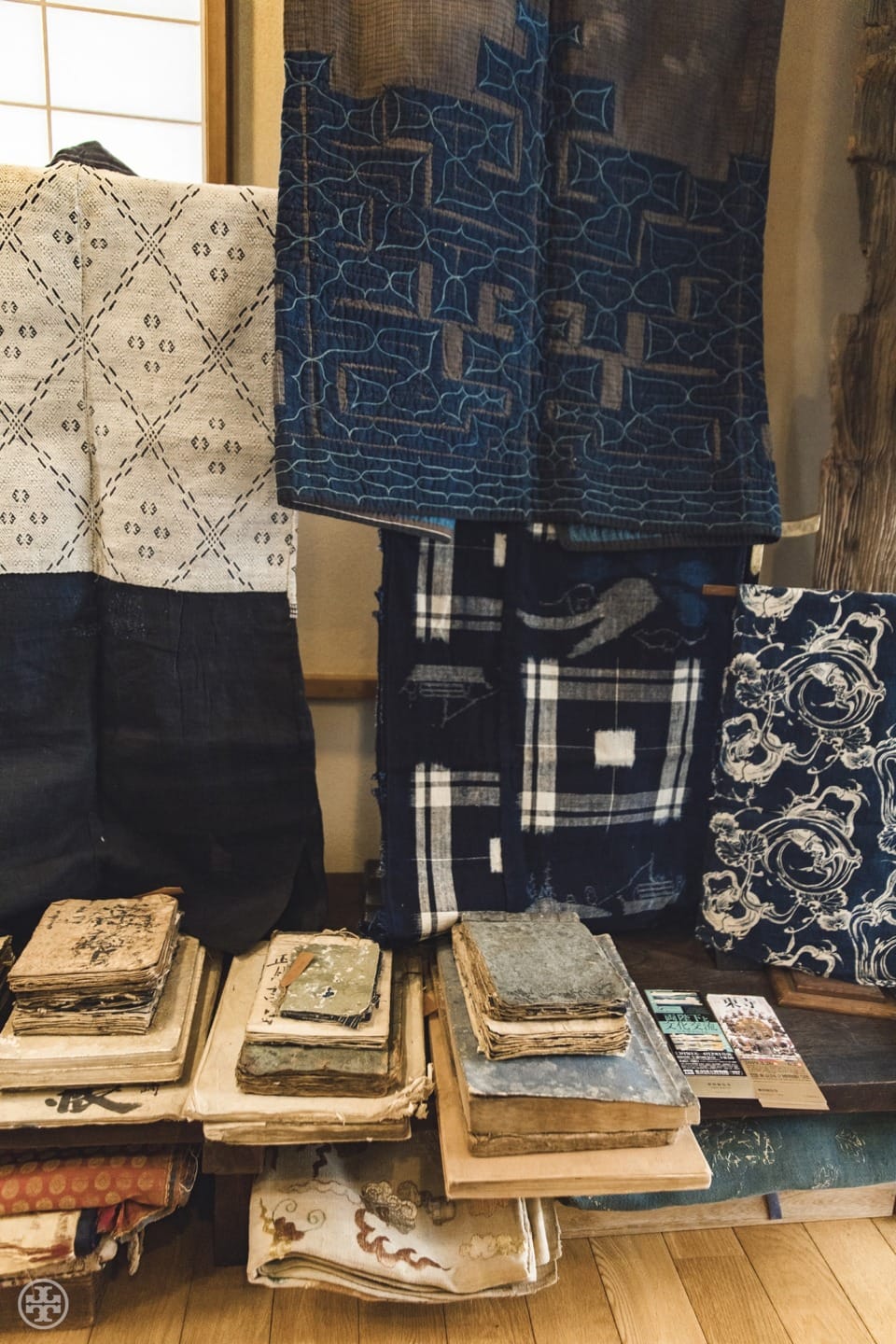 Vintage textiles and old books stacked on a wooden floor, showcasing intricate patterns and textures in a cozy setting.