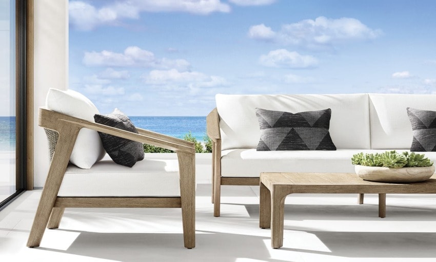 Outdoor patio with white cushions, geometric pillows, wooden furniture, and ocean view in the background.